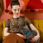 Woman in leopard print top holding a gray cat with a blue bow tie, sitting on a yellow couch against a colorful wall.